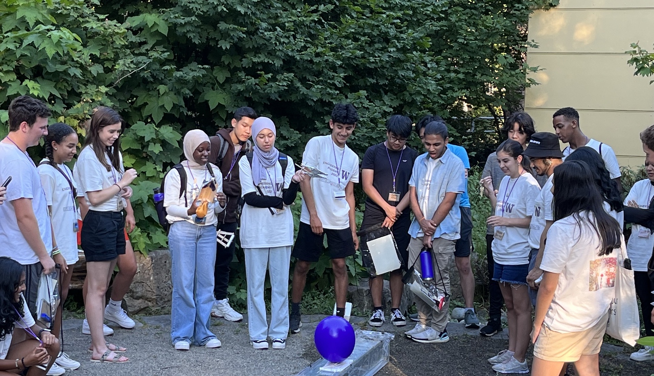 Pre-college students gathering around experimet involving balloon tied to a box