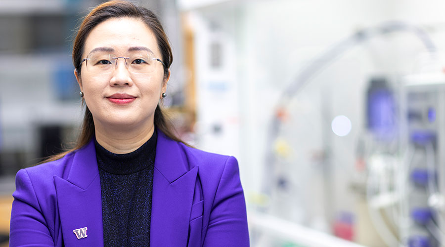 Portrait of Jie Xiao, wearing a purple blazer and UW pin, with her lab in the background.