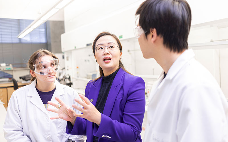 Jie talking to students in her lab. The students are wearing lab coats and eye-protection