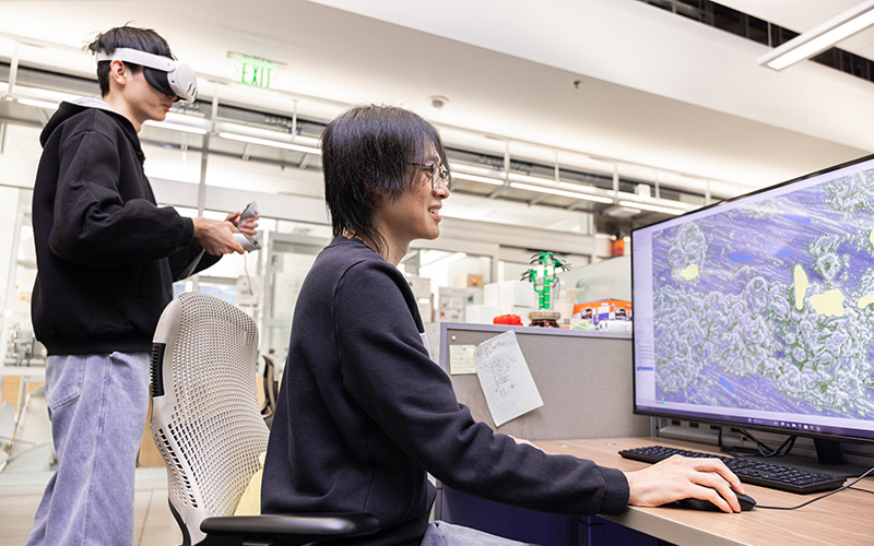 Two students work in front of a computer; one is wearing VR gear and the other is engaging with a screen