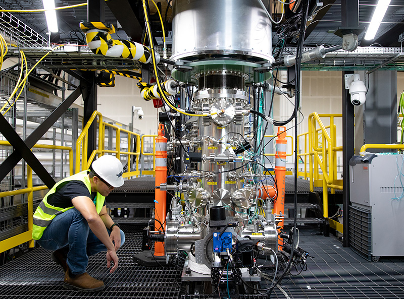 Person wearing high-vis vest and helmet crouches down beside energy fusion device in a factory-like setting