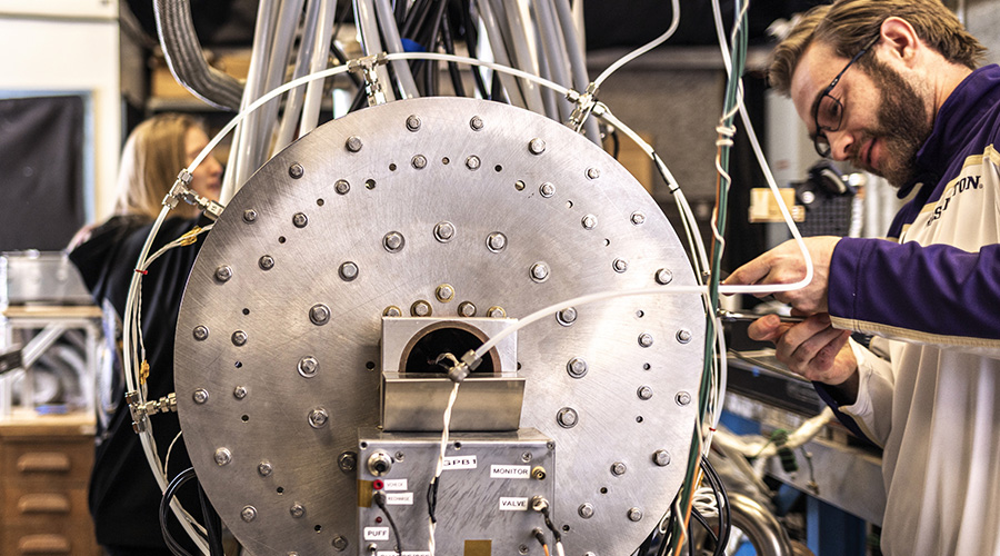 A student wearing UW gear is working on a large machine, with cables and pipes protruding from it.