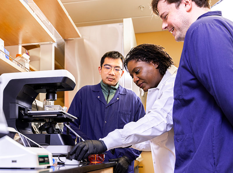 Elizabeth Wayne operating a microscope-looking machine while two students wearing protective gear stand by watching