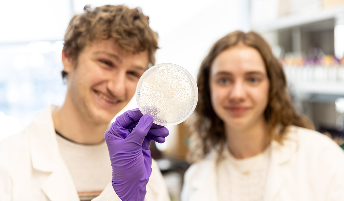 Two students wearing lab coats are in the background, in the foreground, an agar plate being held by a gloved hand