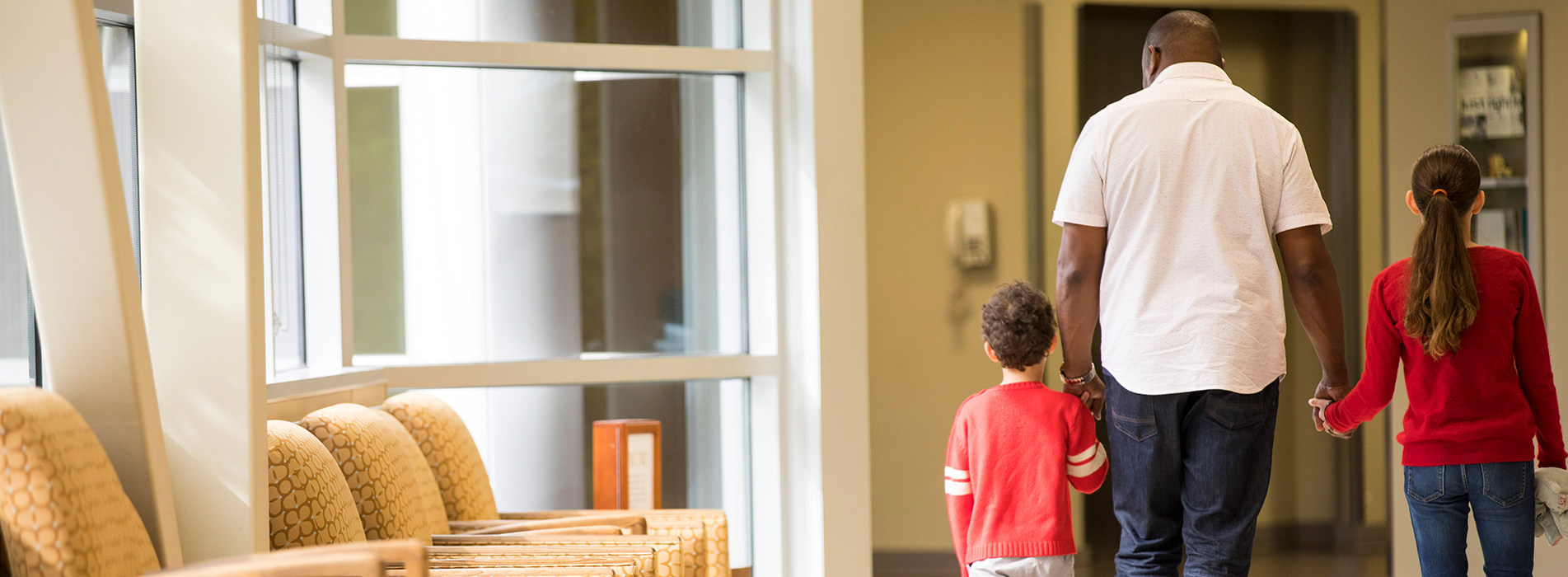 An adult holding the hands of a young boy and girl while walking inside a health-care facility