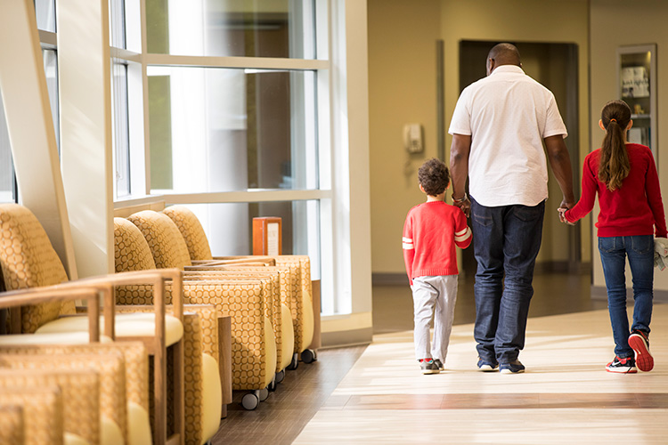 An adult holding the hands of a young boy and girl while walking inside a health-care facility