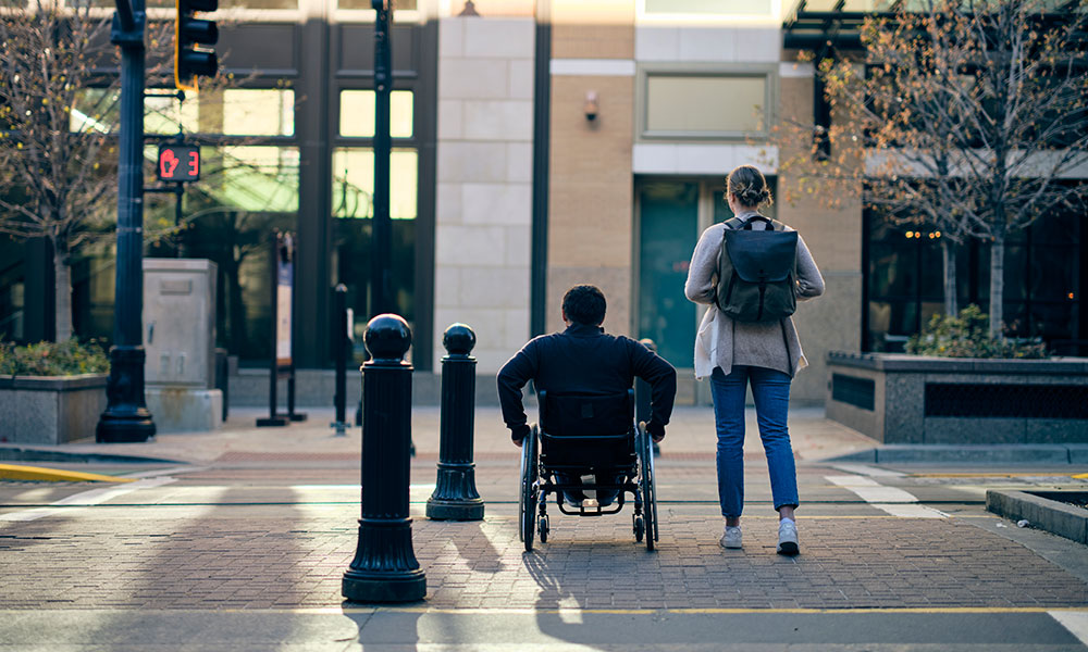 A person in wheelchair and another standing person at a city sidewalk