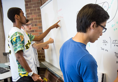 Three students pointing at and writing on a whiteboard