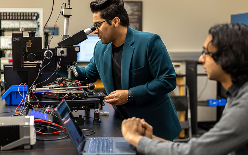 Person wearing suit jacket looking into microscope. One other person observes while working on laptop