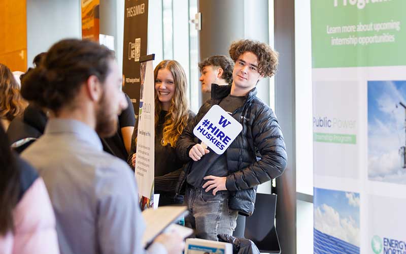 Student holds sign that says "Hire Huskies" in career fair setting