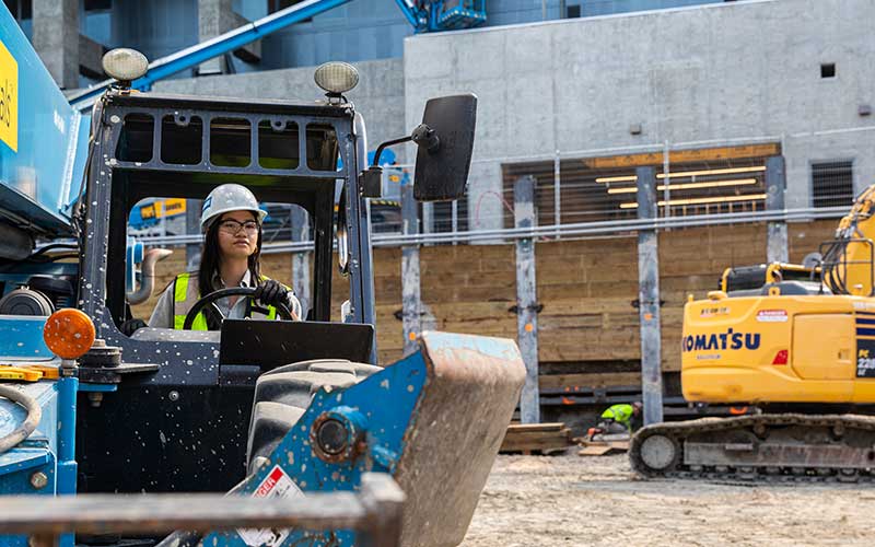 Student wearing hard hat and high visibility vest driving heavy machinery on constructionsite