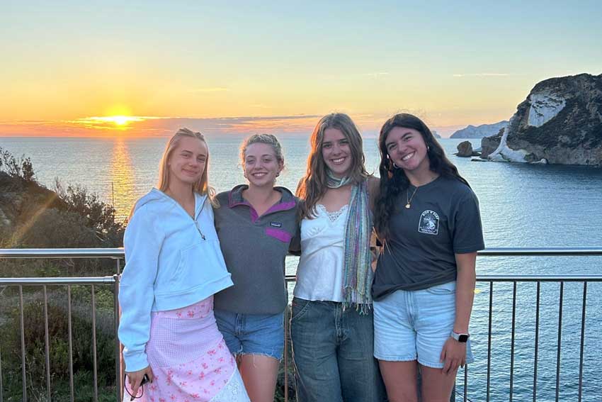 Four students stand in front of a picturesque coastal view of Italy as the sun sets over the sea