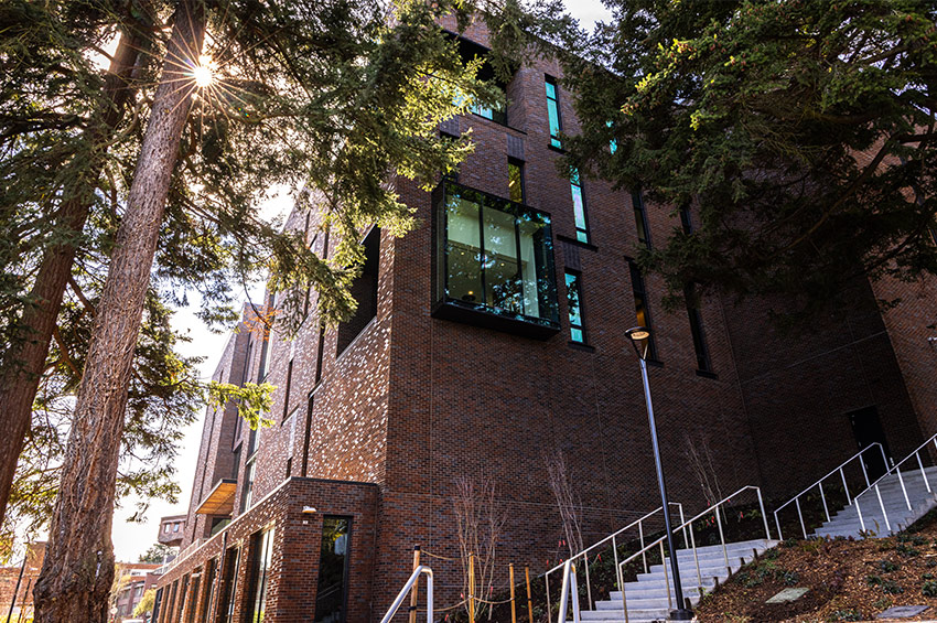 A brick building with protruding glass section, surrounded by trees and sunlight.