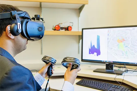 Professor Akshay Gadre wearing an AR/VR headset, holding controllers, while sitting in front of a computer screen showing a blueprint map of the UW campus