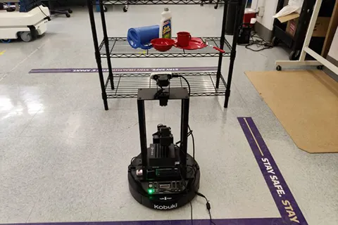 A robot in front of a shelf. The shelf contains the following items: a pitcher on its side, a bowl in front of a bottle of Soft Scrub, a mug on a plate and a spoon balanced on the plate