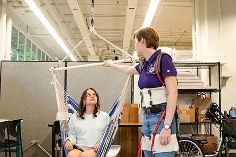 One person is sitting in a hammock chair, while another person holds part of the frame structure