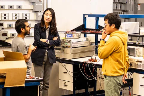 UW ECE Assistant Professor Jungwon Choi standing in between two graduate students with her arms crossed, in conversation. The room is full of boxes, wires, and electronic equipment.