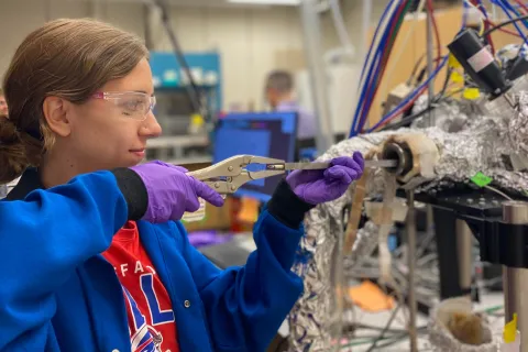 A woman in a blue lab coat and safety goggles holds an instrument to load a sample into a reactor in the lab