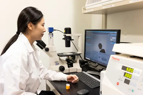 Person in a lab coat working with a microscope and computer in a laboratory.