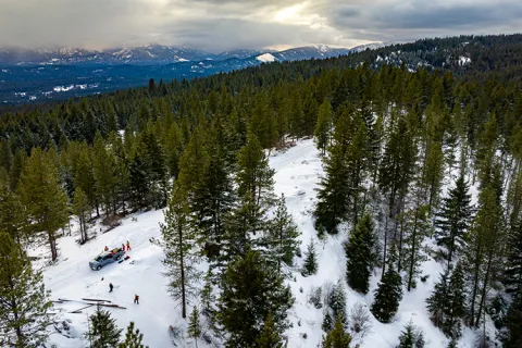 An aerial photo of a snowy forest with a mountain range in the background. In the foreground, several small figures stand next to a pickup truck.
