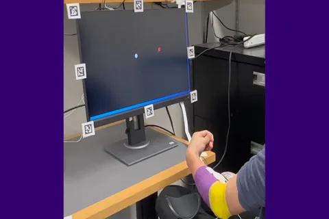 A person's arm, wrapped with purple and yellow tape, gestures in front of a black monitor displaying a red and blue dot.