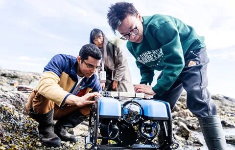 Three people on a rocky shore examine a small blue underwater drone. Two kneel beside it, focusing intently, while the third person stands in the background. They are surrounded by seaweed under a cloudy sky.