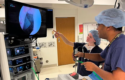 Nicole Gunderson and Waleed Abuzeid conduct an experiment in a hospital setting. Gunderson points at a screen showing a video of a sinus model while Abuzeid holds a probe inside a sinus model.
