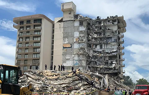 A large condominium that has partially collapsed, and people with helmets amidst the rubble.