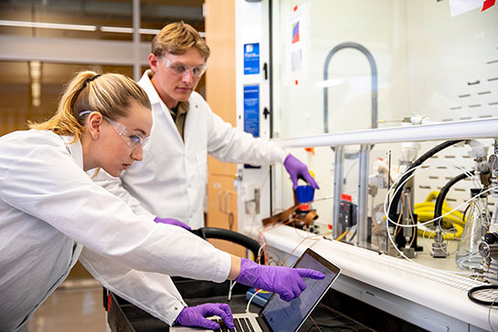 two researchers in lab coats and one points at a laptop monitor