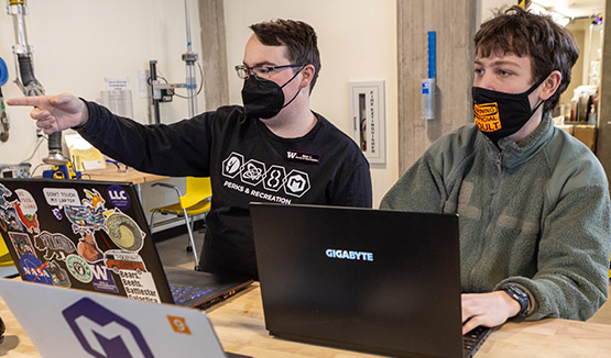 Two students sitting next to each other in front of laptops as one student points to an object outside of the photo