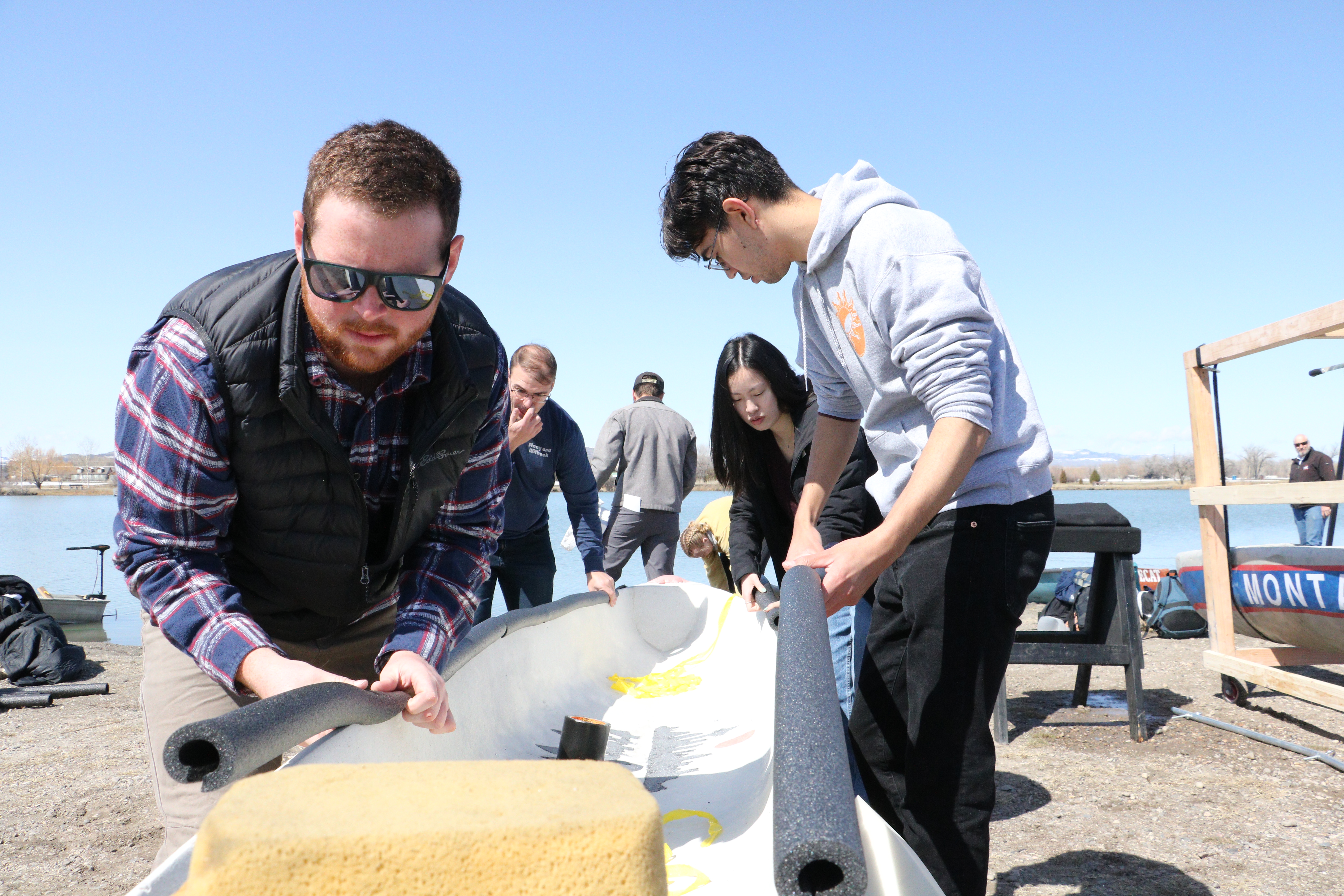 Two students stand on either side of a canoe and fasten foam insulation to the sides