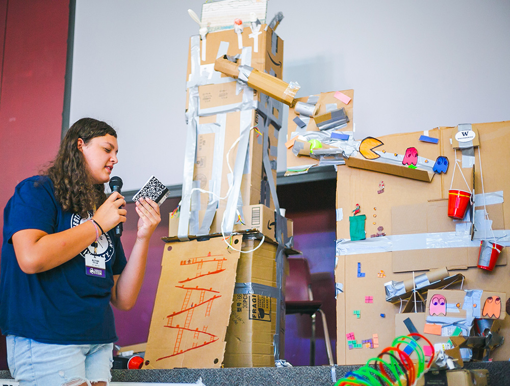 Aliyah Taylor talking into a mic while looking at a small notebook in front of a Rube Goldberg inspired machine built out of cardboard