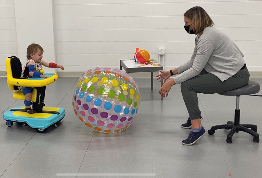 A toddler-aged child in a ride-on toy gaining mobility to explore other toys, accompanied by a researcher.