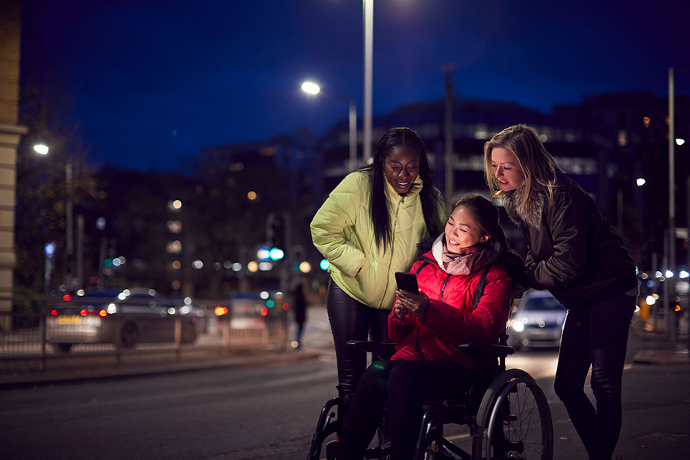 A person sitting in a wheelchair looking at a phone while two people are looking over her shoulder at the phone