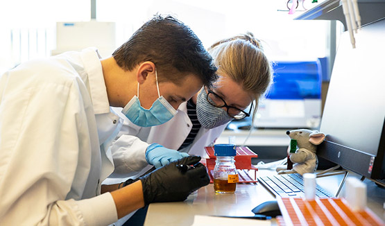 Two people working in a lab setting, looking at a subtance in a vial while wearing face masks