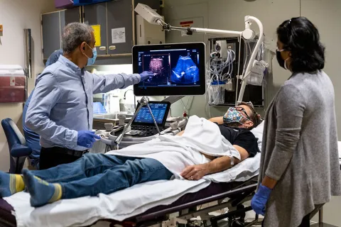 Mike Averkiou pointing at a monitor that shows a contrast-enhanced ultrasound of Lutz's liver while Barry Lutz is lying in bed with Dr. Manjiri Dighe standing at his left