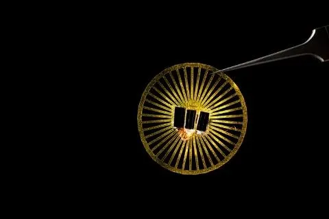 Tweezers holding a circular device with solar panels on it. It's illuminated against a black background.