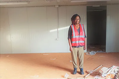 Judy Khalamwa standing at an indoor construction site wearing an orange safety vest 