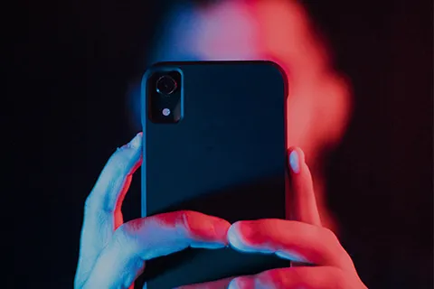 Closeup of person's fingers grasping mobile phone with camera facing outward in eerie red and blue light, person's face and hair are blurred behind the phone against a dark background