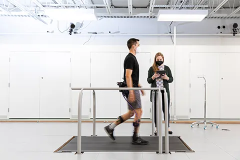 Man walking on a treadmill wearing an exoskeleton device while a female researcher stands next to him monitoring his activity on a tablet