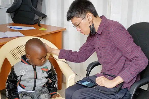 A child sits in a chair and a person sits next to them. The person is holding a probe to the child's ear. The probe is connected to a smartphone on the person's lap.