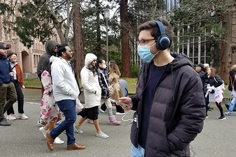 A man wearing a surgical mask and headphones walks through the University of Washington campus while holding a smartphone. People walk behind him.