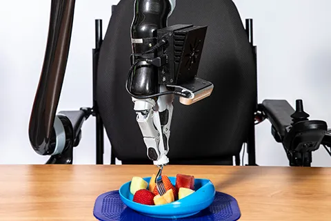 An assistive-feeding robotic arm attached to a wheelchair uses a fork to stab a piece of fruit on a plate among other fruits.