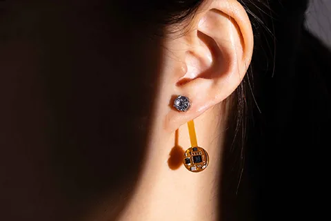 The temperature sensing earring is shown attached to a person’s ear, touching the earlobe is a gemstone while dangling a few centimeters below is a small circular circuit board.