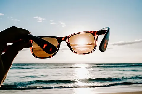 A person's hand is holding a pair of sunglasses against the sky so that the sun shines through one lens while illuminating ocean waves.