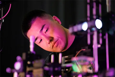 Bingzhao Li working with lab equipment. The room is dark and the lab equipment is giving off a purple glow from lasers used in Li's work.