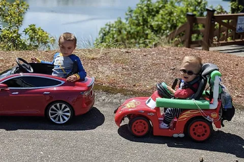 Two children ride in small toy cars, one of which has an adapted steering wheel to make it accessible for the child to use.