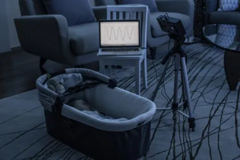 A baby is asleep while a smart speaker prototype monitors its breathing. The breathing waveform is shown on a computer screen nearby. 