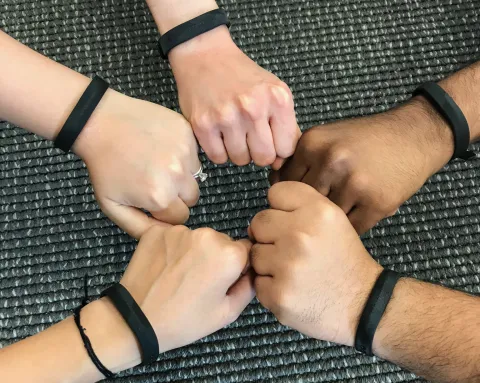 Five hands making fists in a circle. All arms have black Fitbit trackers on them.