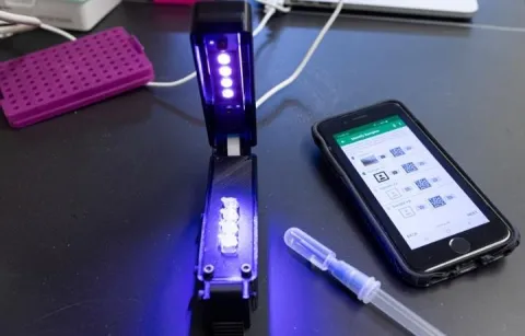 The Harmony COVID-19 test includes a small, inexpensive detector (left) that can processes four reaction tubes — shown in the detector — and a sample collection device (center). Results are displayed on a smartphone (right).